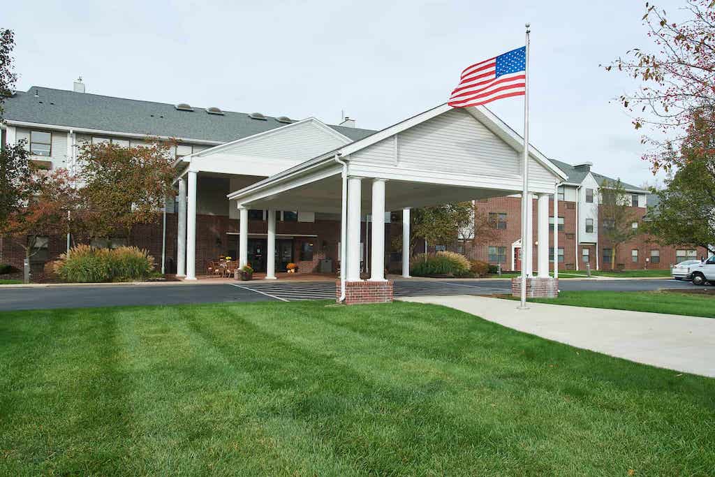 Front of senior living community with a covered driveway, lush grass lawn, and a pole flying the United States flag.